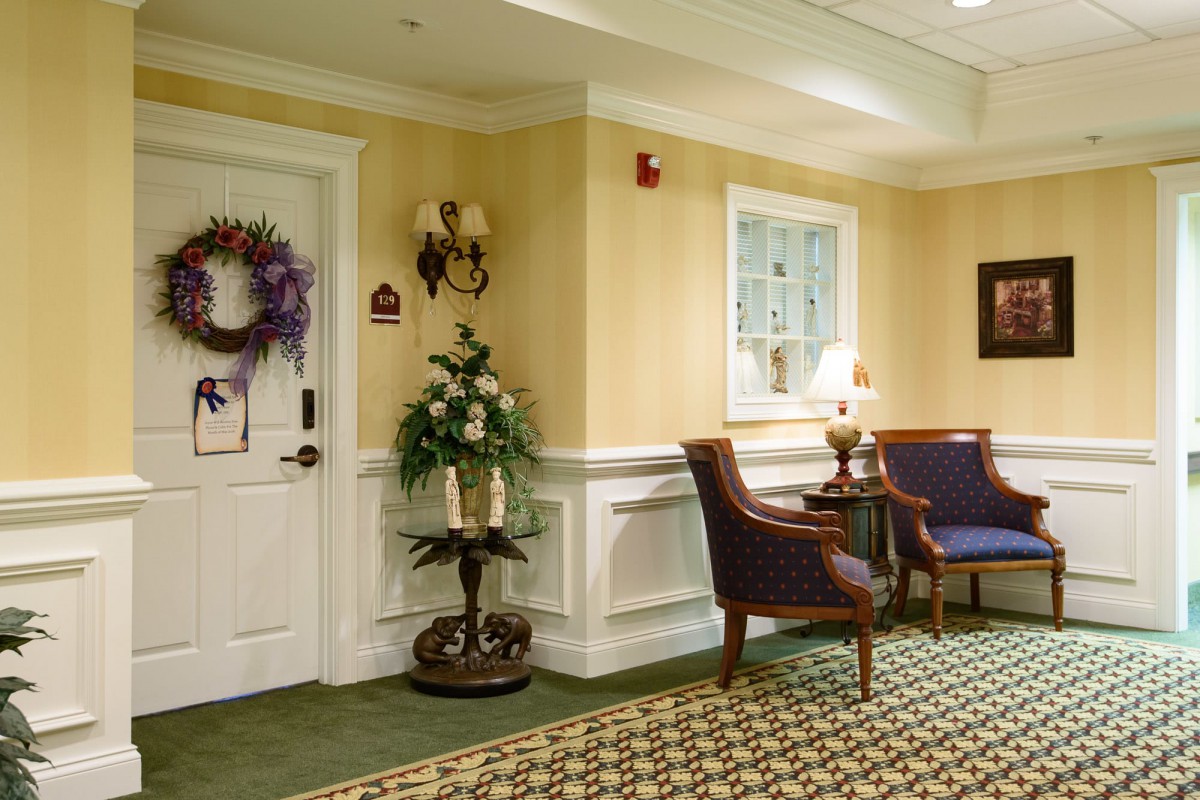 A warmly decorated hallway with a seating area, featuring chairs, a small table with a lamp, and decorative elements including a wreath on the door.