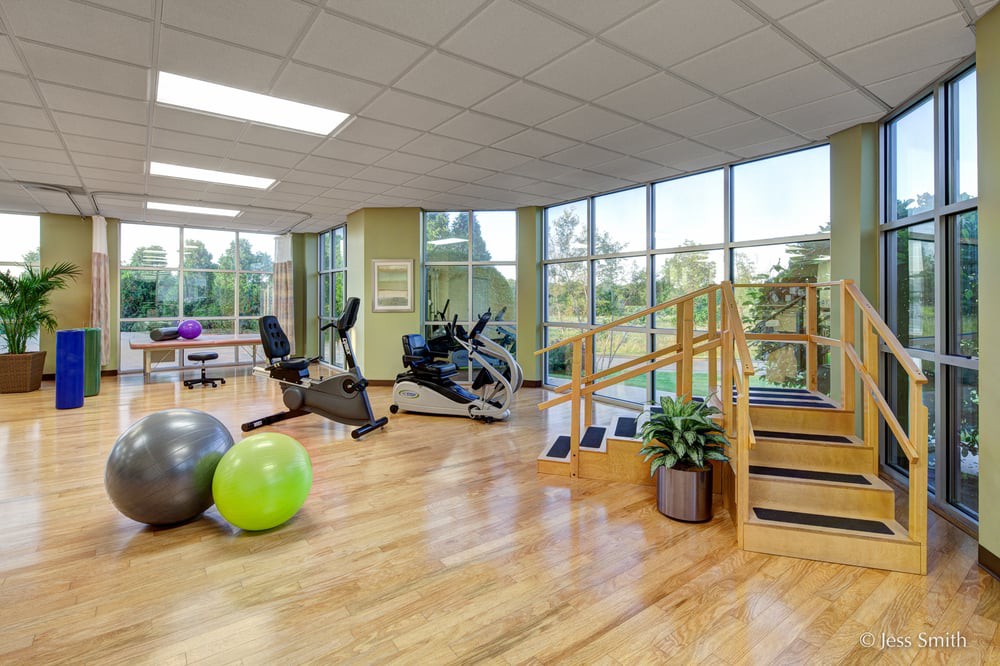 A spacious gym with exercise equipment, including an elliptical machine, exercise balls, and stairs, featuring large windows and wooden floors.