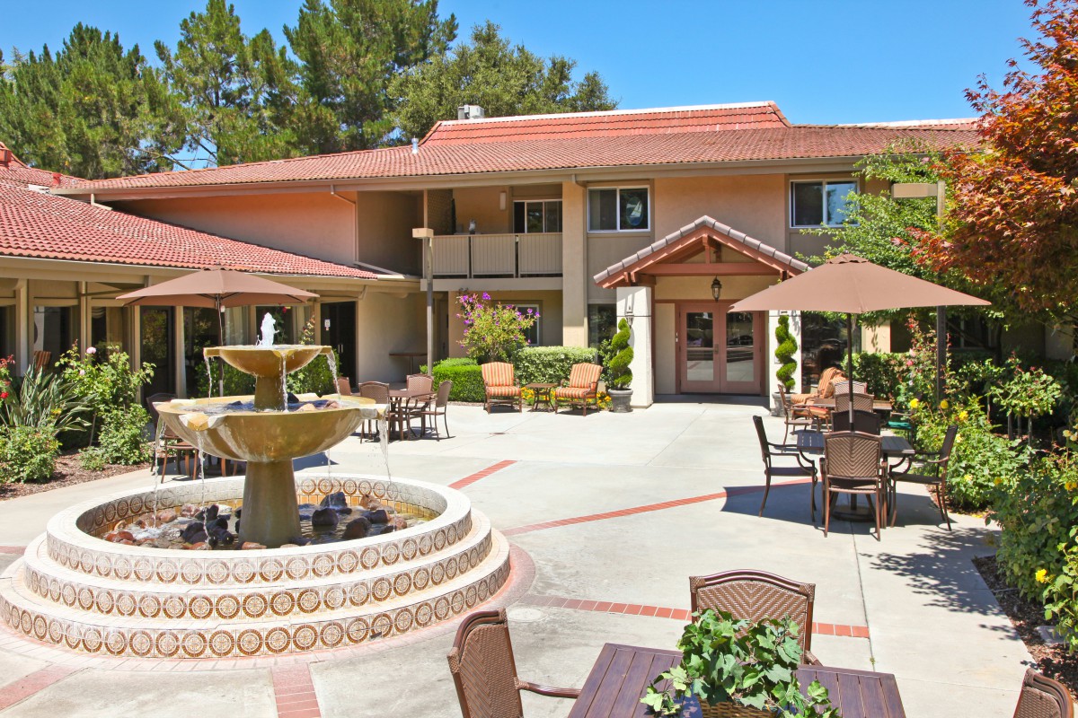 A sunny courtyard with a fountain and outdoor seating in front of a two-story building.