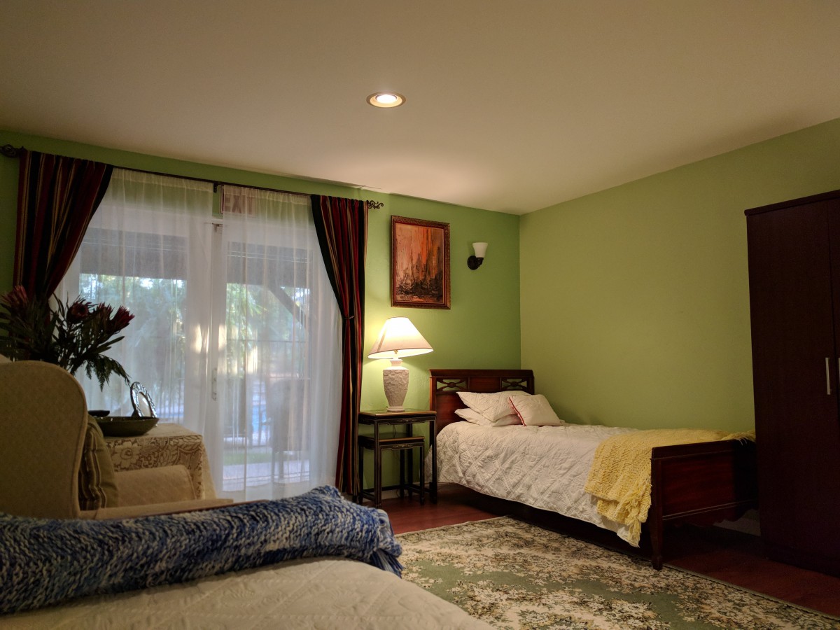A cozy bedroom with a single bed, accented by green walls, a lamp on a bedside table, and large windows with curtains.