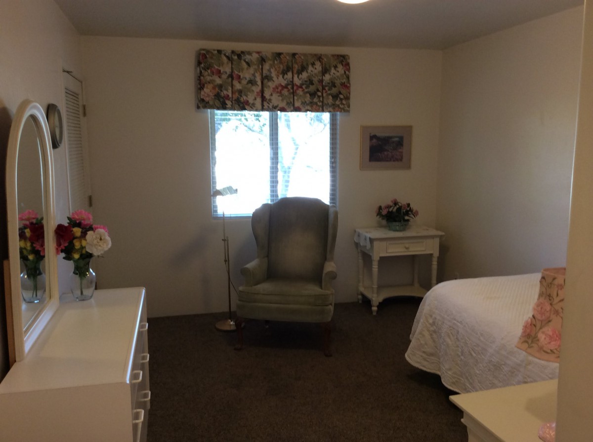 A cozy bedroom featuring a green armchair by the window, a bedside table with flowers, and a neatly made bed.