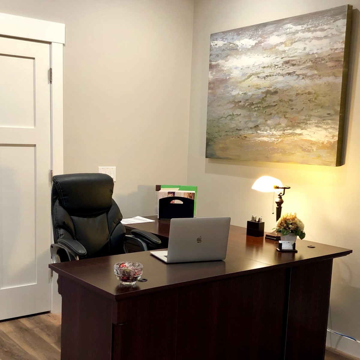 A neat and organized office space featuring a desk with a laptop, chair, lamp, and artwork on the wall.