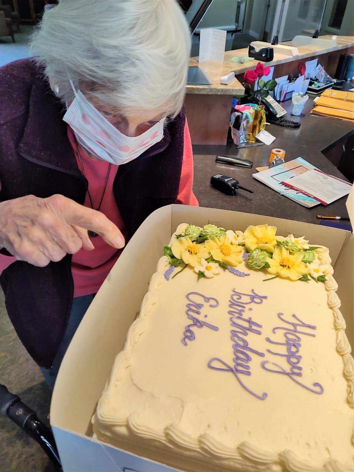 An elderly person wearing a mask admires a birthday cake with yellow flowers and the message 'Happy Birthday Erika'.
