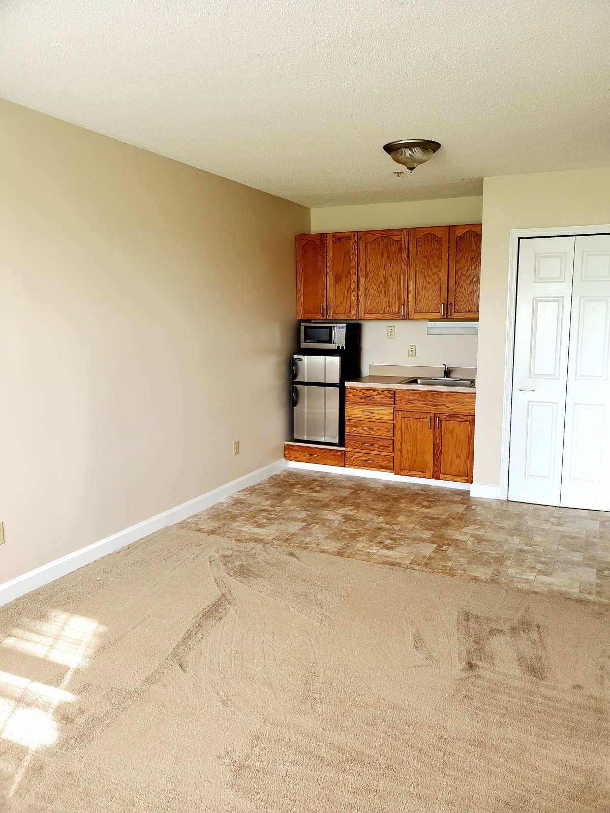 An empty apartment with a small kitchenette and beige carpet.