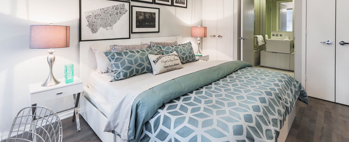 A cozy bedroom with geometric-patterned bedding and decorative pillows, complemented by framed wall art and a soft-lit lampshade.