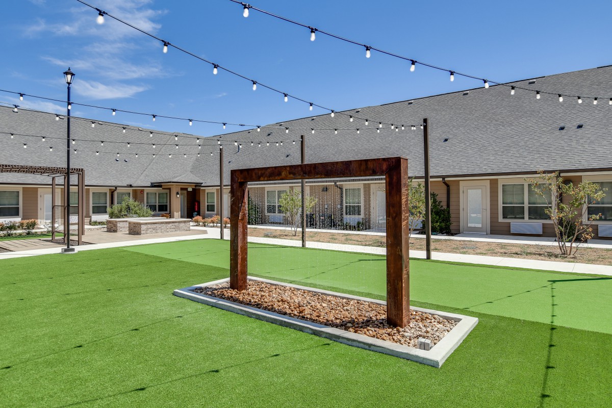 A modern courtyard featuring a wooden structure, artificial grass, and string lights on a sunny day.