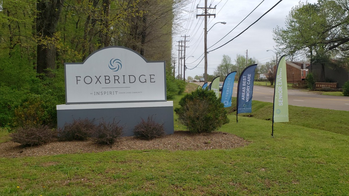 The Foxbridge entrance sign is situated along a roadside with greenery and promotional flags.