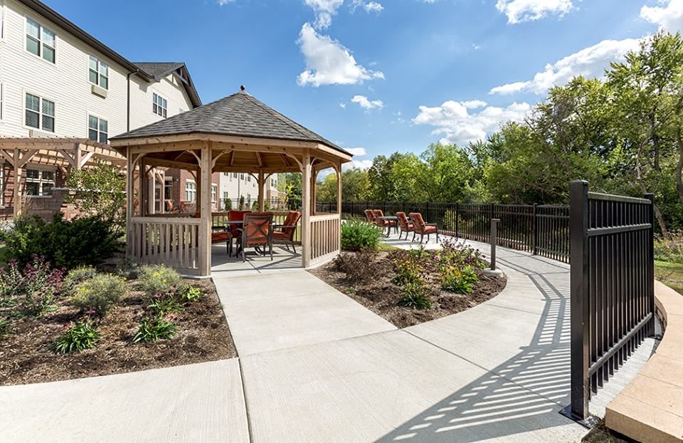 A scenic outdoor area featuring a gazebo with seating, surrounded by a landscaped garden and pathway under a clear blue sky.
