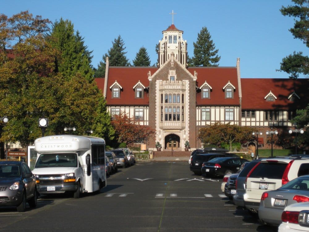 A historic building with a red roof and tower, viewed from a parking lot bordered by trees.