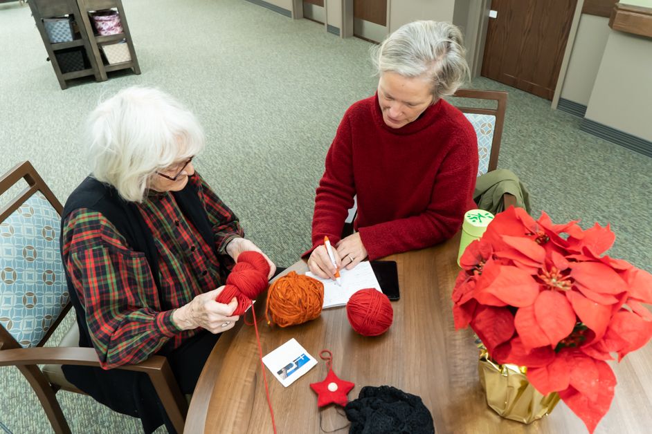 Two elderly women sitting at a table, engaged in knitting with various colored yarns.