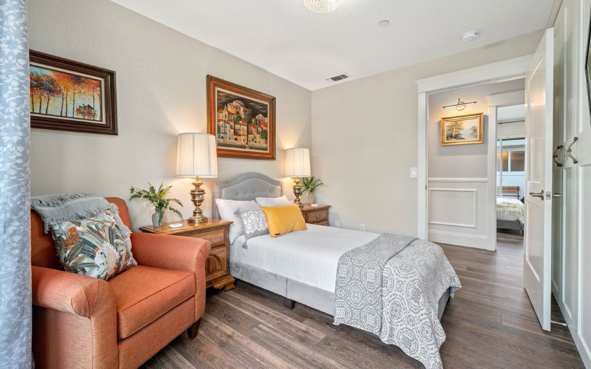A cozy bedroom featuring a single bed with decorative pillows, a pair of lamps on wooden side tables, an armchair, and artwork on the wall.