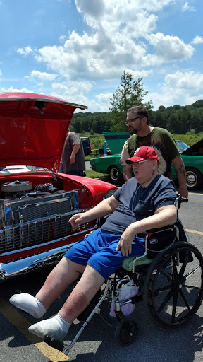 A man in a wheelchair enjoys the sights at a local car show, with a vintage red car on display.