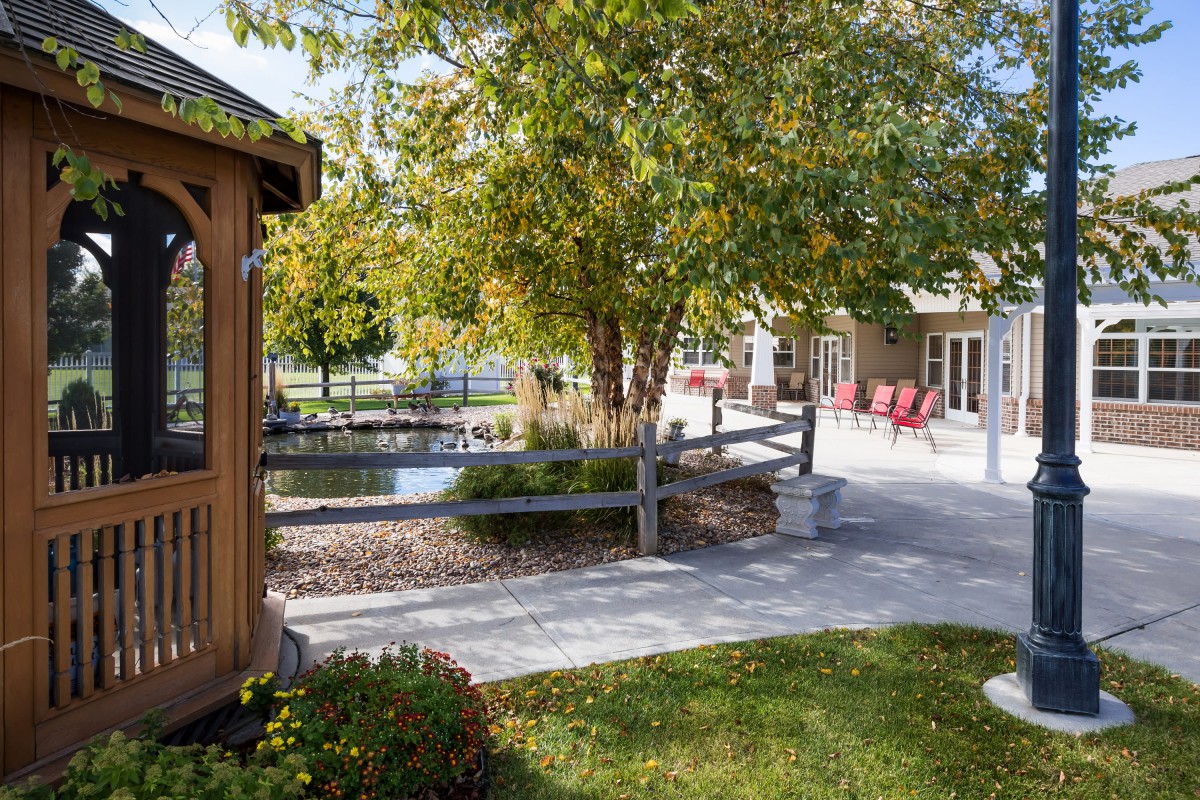 A serene outdoor garden with a pathway, surrounded by trees and a wooden fence, leading to a seating area.