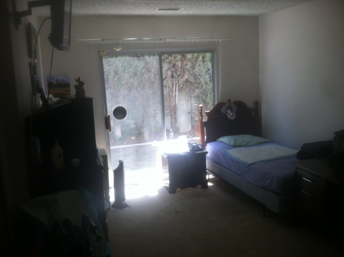 A dimly lit bedroom with a single bed and sunlight streaming through a large window.