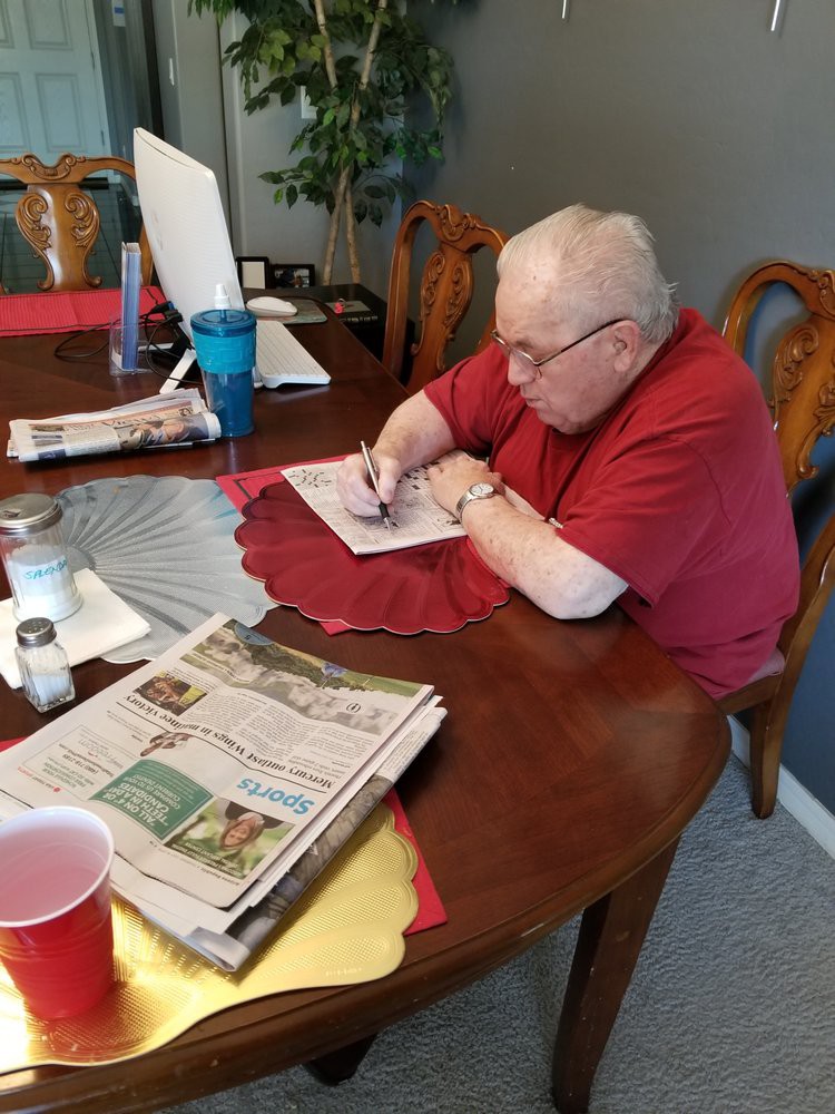 An elderly person sitting at a table, focused on writing with newspapers nearby.