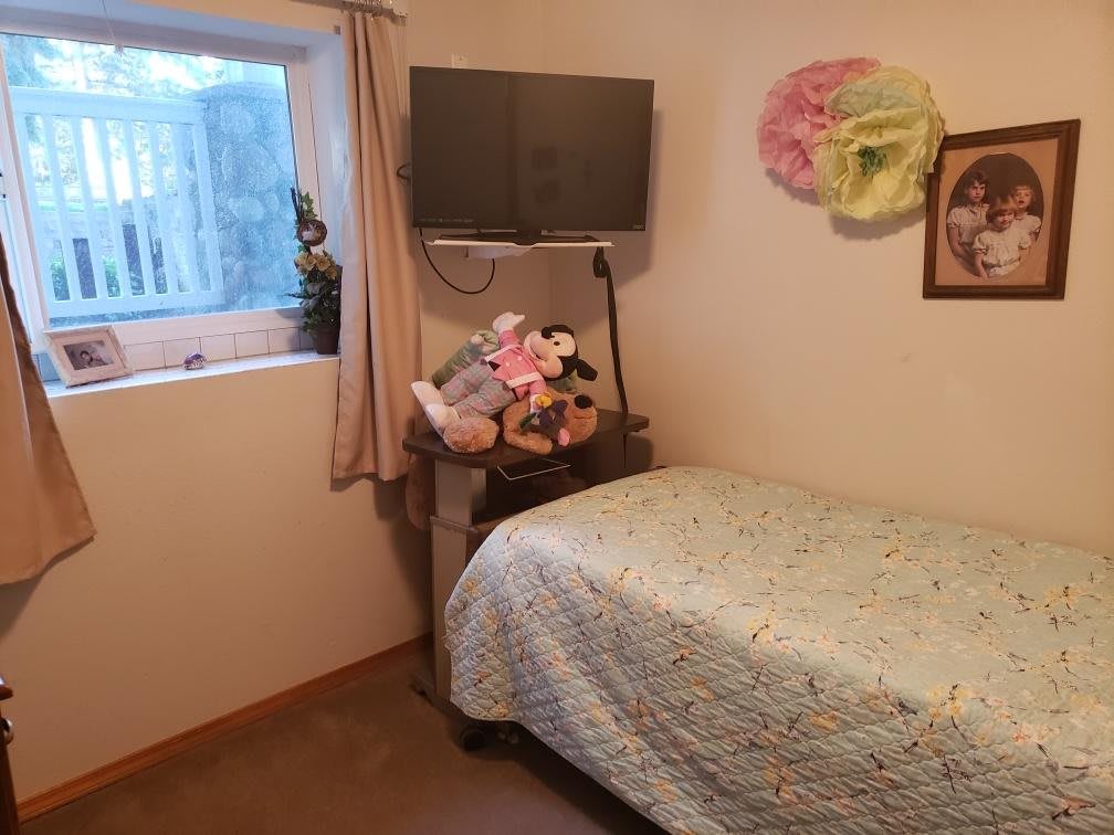 A cozy bedroom with a single bed, a quilt cover, and a small collection of stuffed animals on a shelf under a wall-mounted TV. The room features a window with curtains and decorative items on the walls.