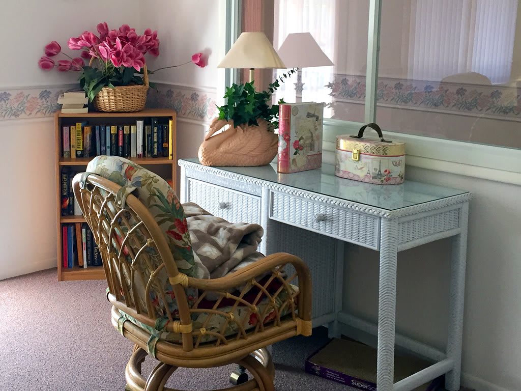 A cozy home office setup features a wicker desk and chair, with a lamp, books, and a basket of flowers creating a welcoming atmosphere.