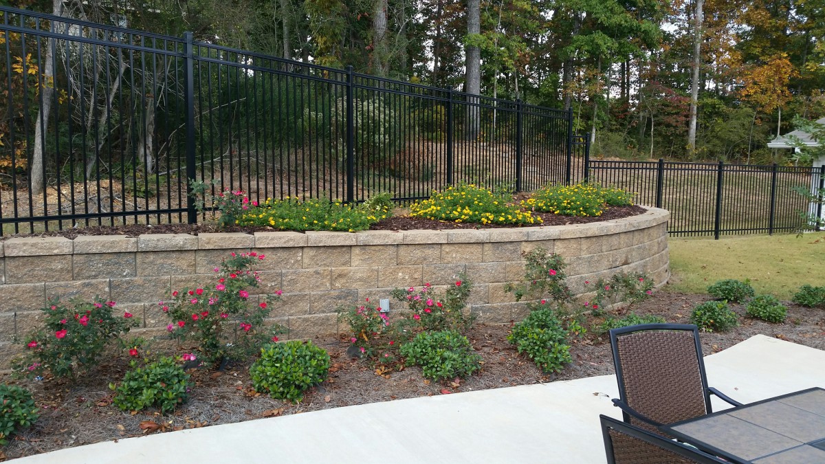 A landscaped garden with a stone retaining wall, metal fence, and neatly arranged flowers.