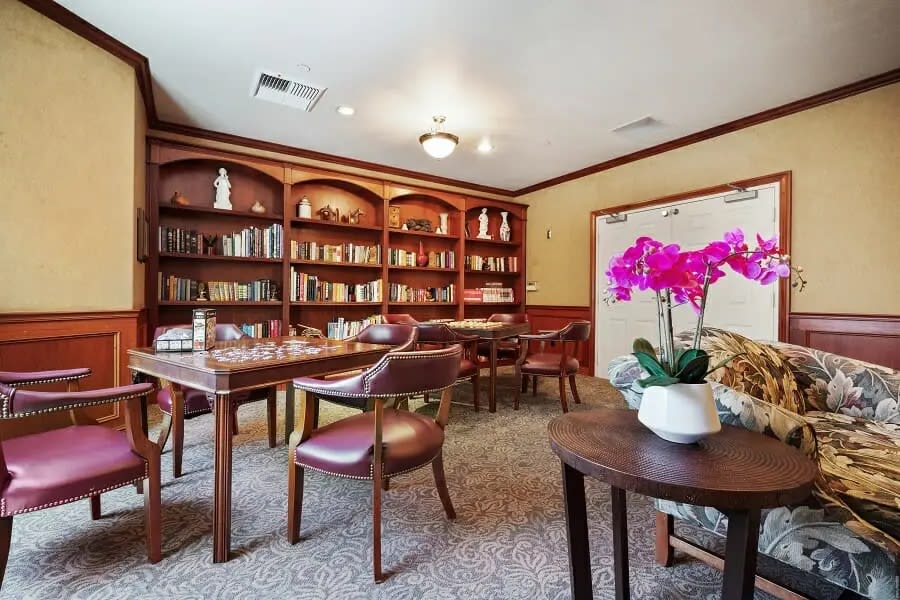 A cozy library room with wooden bookshelves, tables, and chairs, featuring a vibrant potted orchid on one table.