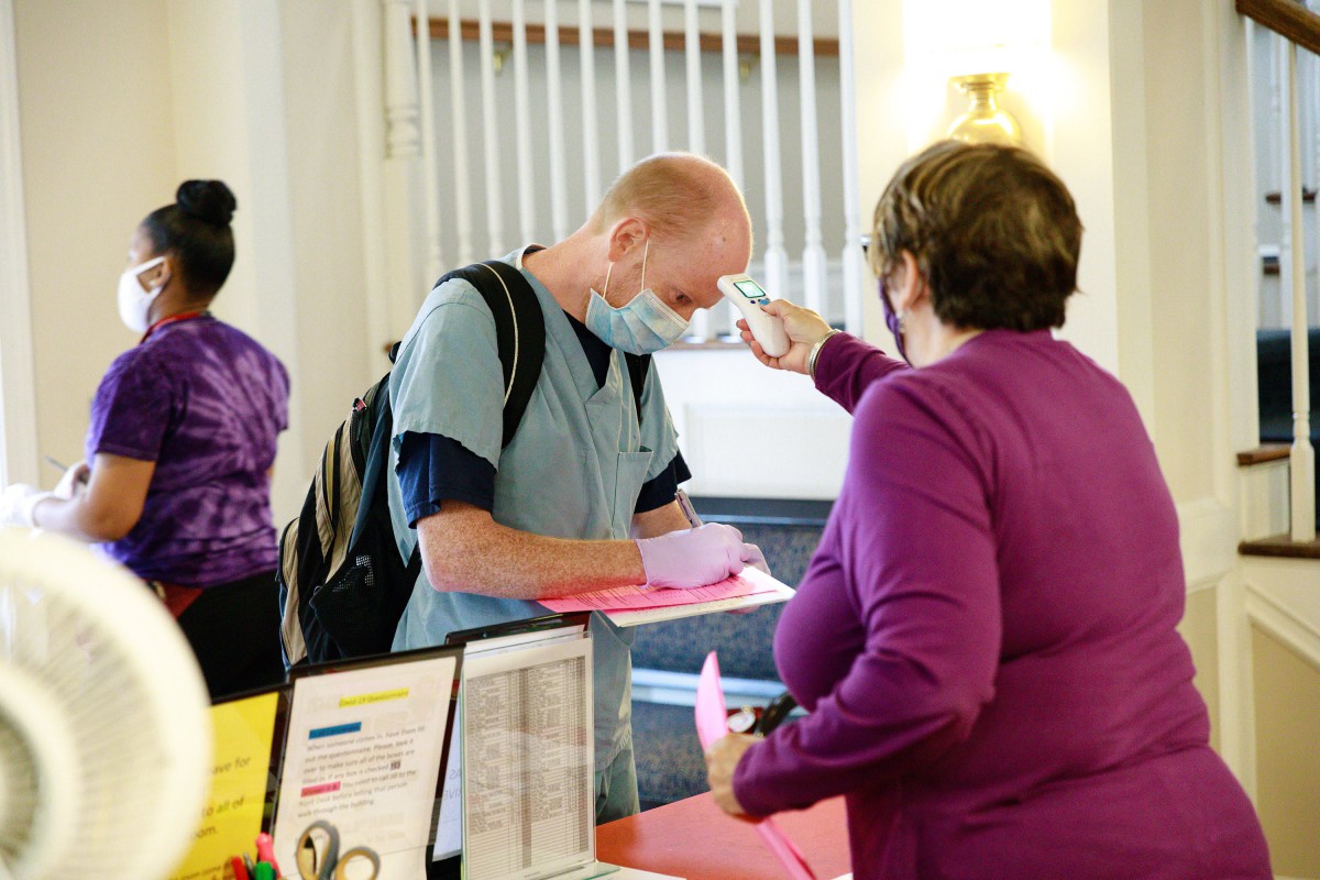 A healthcare worker undergoes a temperature check at a check-in desk while wearing a mask.