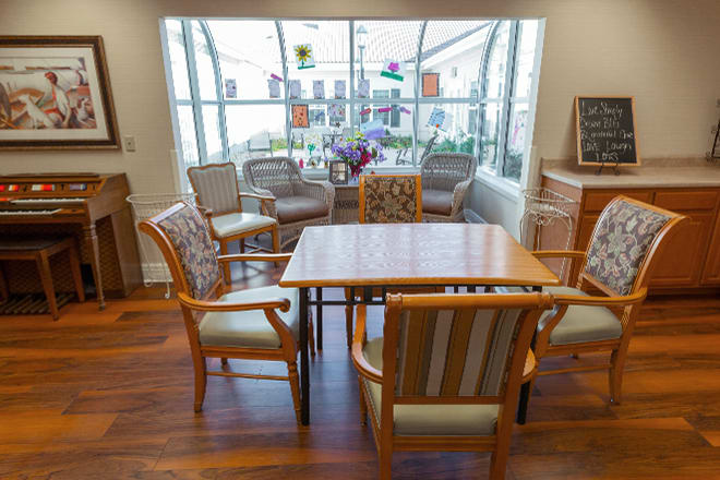 A cozy living room setup with a wooden table surrounded by chairs, decorated with flowers and colorful window ornaments.
