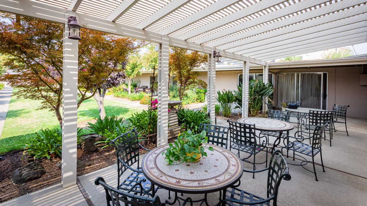 A serene patio area with a table and chairs under a pergola, surrounded by lush garden greenery.