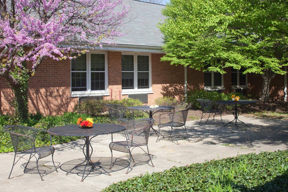 An outdoor patio with tables and chairs surrounded by vibrant trees and a brick building in the background.