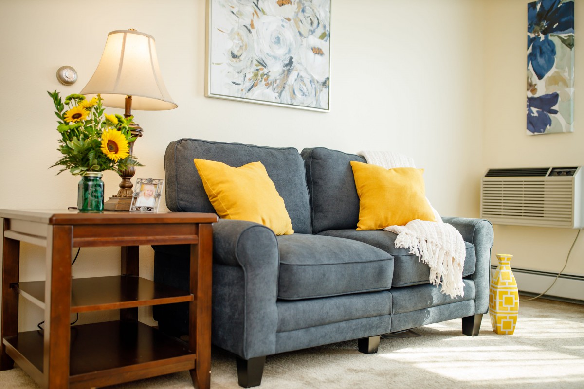 A cozy living room features a blue sofa adorned with yellow pillows, a wooden side table with a lamp, and a sunflower in a vase.