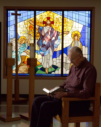 An elderly man engages in quiet reflection while reading in front of a beautifully crafted stained glass window.