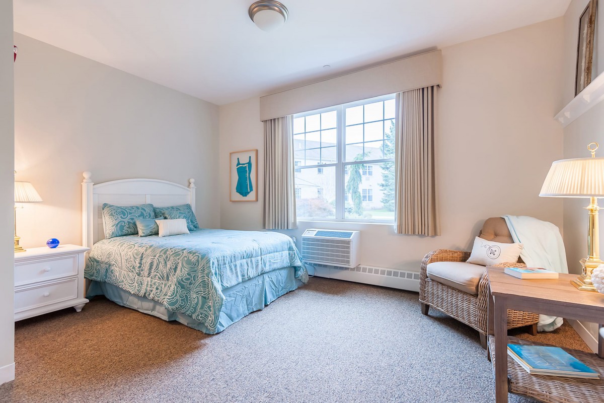 A cozy bedroom with a neatly made bed, large window, and comfortable chair.