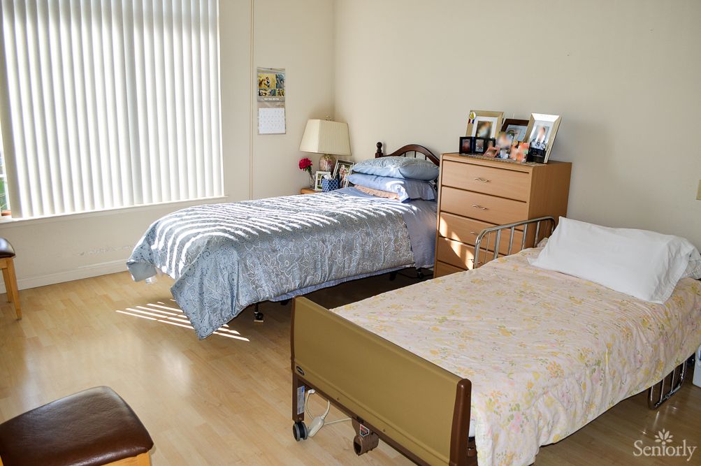 A bedroom featuring two beds, one resembling a hospital bed, with a dresser and window blinds.