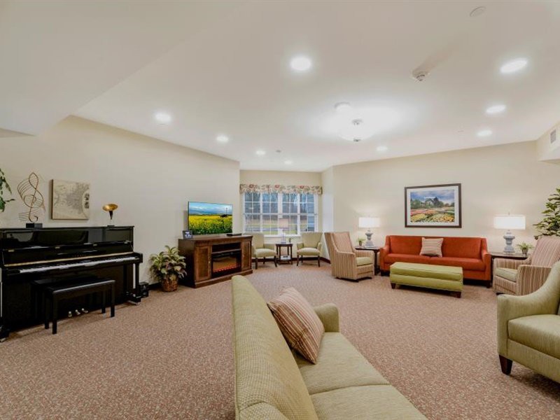 A spacious and well-lit living room featuring a piano, television, multiple sofas, armchairs, and a large window.