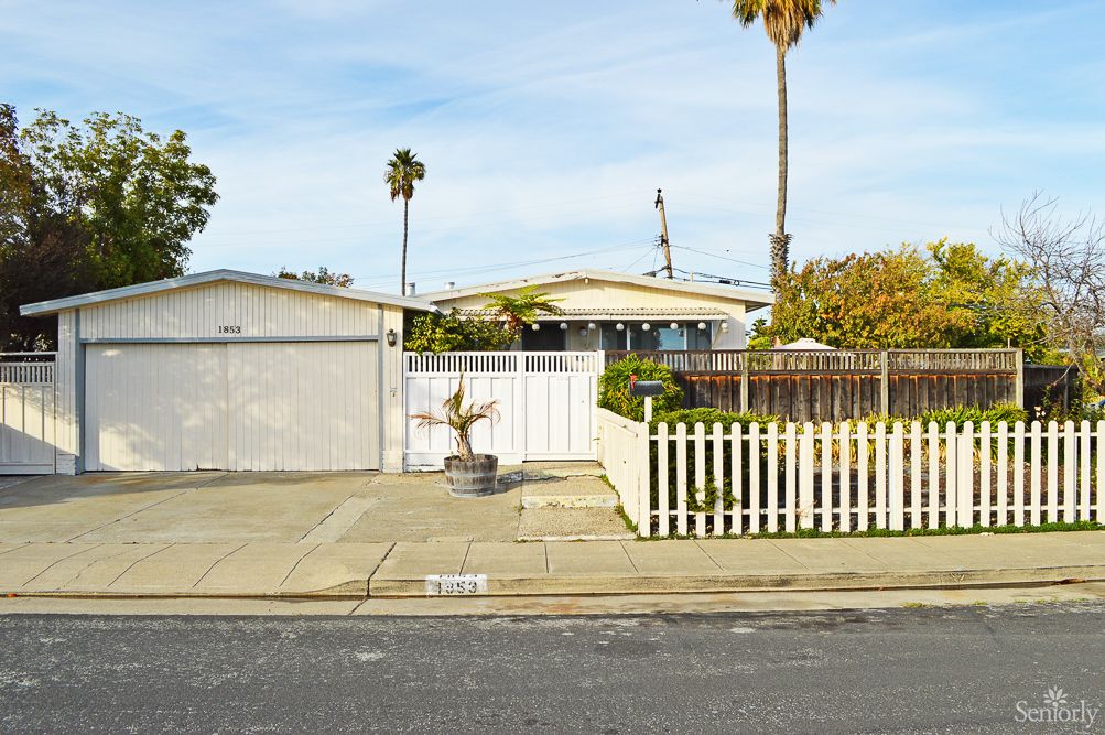 A suburban house with a picket fence and driveway, featuring palm trees in the background.