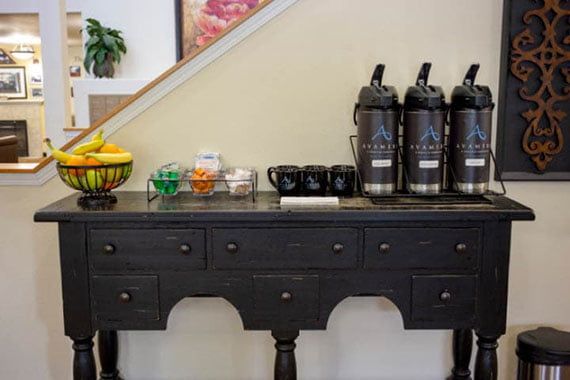 A coffee station with dispensers, mugs, and a fruit basket on a black table.