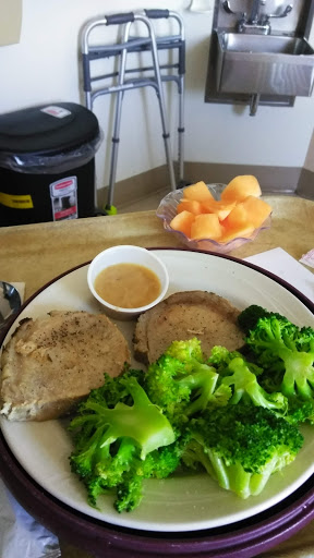 A plate of sliced cooked meat served with broccoli and a small bowl of dipping sauce, accompanied by a side of fresh cantaloupe.