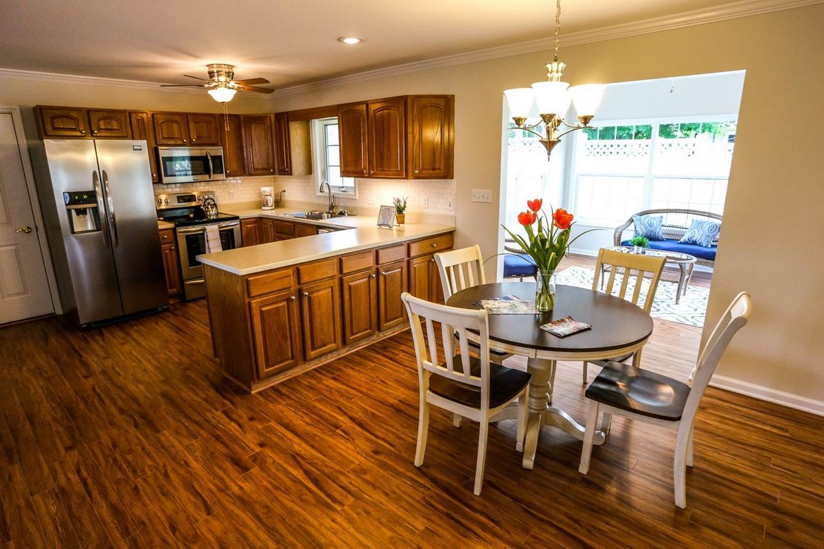 A cozy kitchen and dining area with wooden cabinets and flooring, featuring a table with chairs and a vase of flowers.