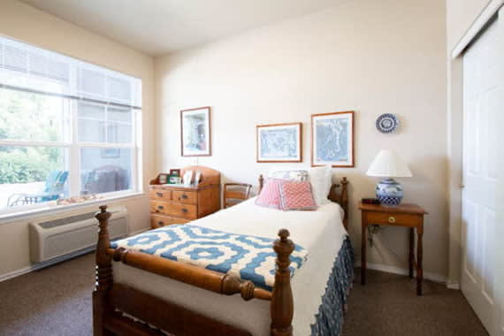 A cozy bedroom with a single wooden bed, decorated with a blue and white quilt, framed art on the walls, and natural light streaming through the window.