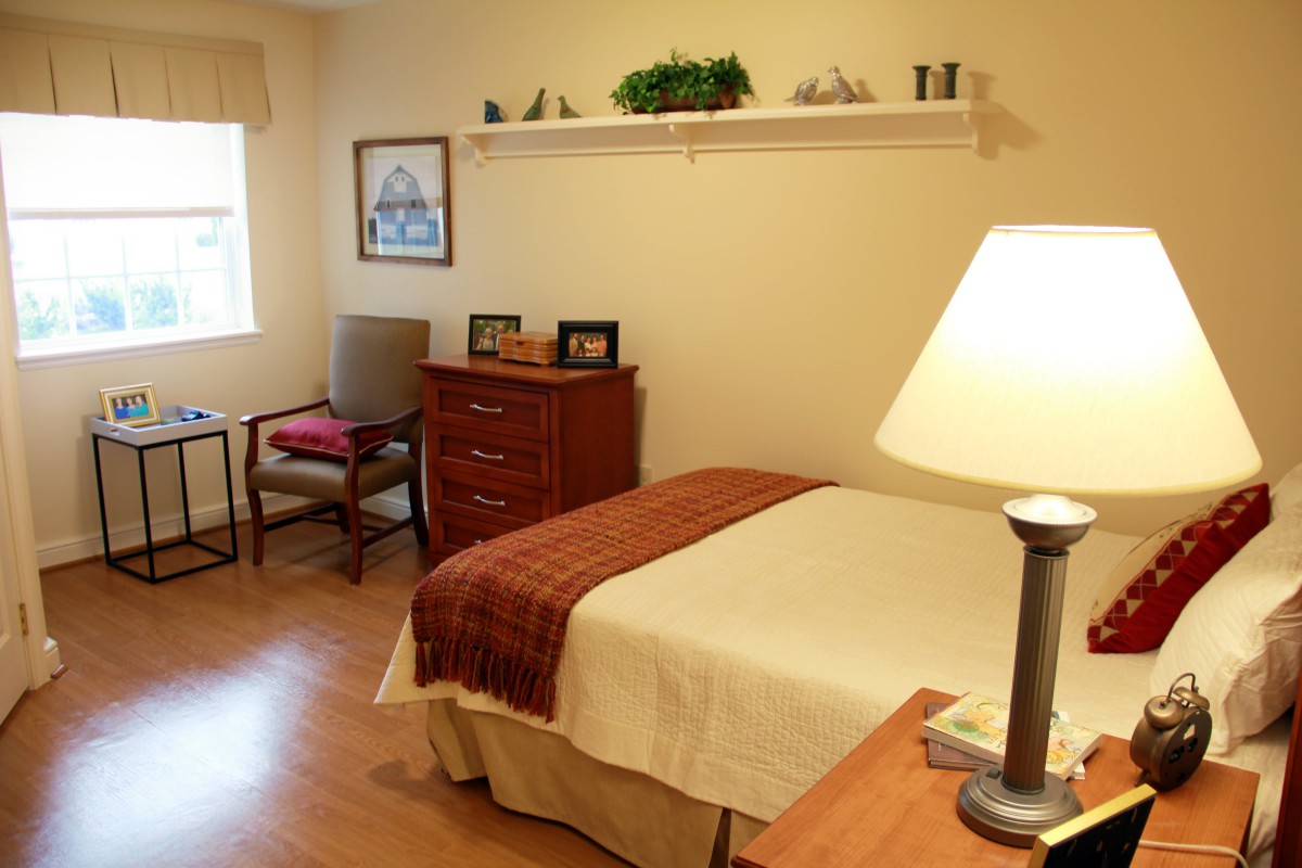 A cozy bedroom with a neatly made bed, a wooden dresser, and a lamp on the side table.