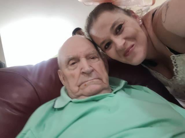 A woman takes a selfie with an elderly man sitting on a couch.