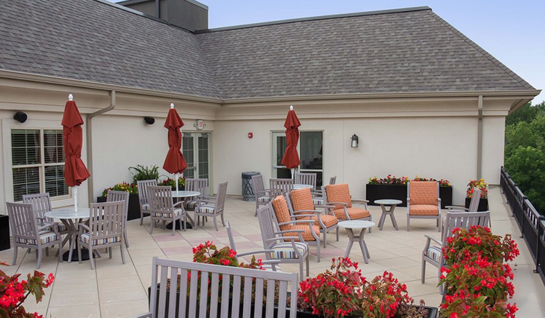 A rooftop patio featuring orange-cushioned chairs, tables with red umbrellas, and vibrant flower arrangements.