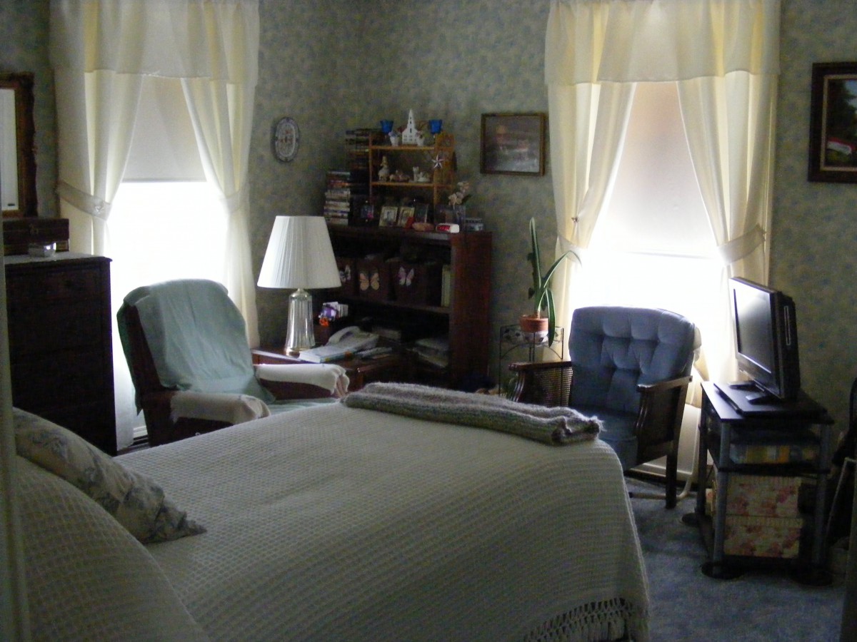 A cozy bedroom with a neatly made bed, chairs, and shelves filled with books under soft lighting.