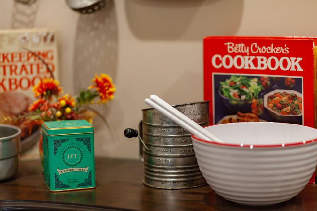 A vintage kitchen setup featuring a cookbook, mixing bowl with utensils, and decorative flowers.