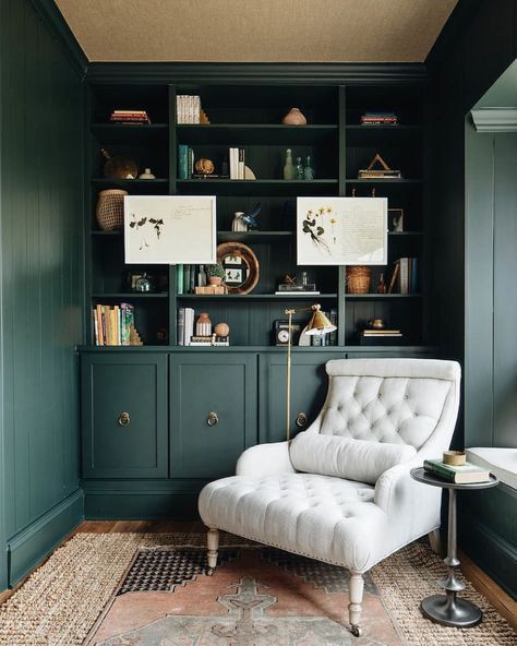 A cozy reading nook with a comfortable armchair and dark green built-in bookshelves adorned with decor and artwork.