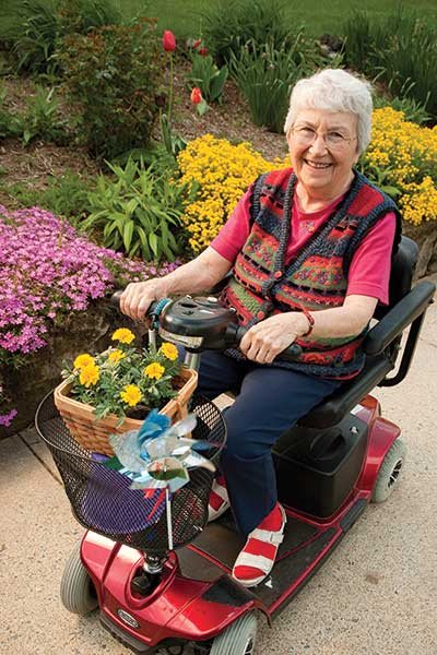 An elderly woman enjoying a ride on her mobility scooter, surrounded by blooming flowers.
