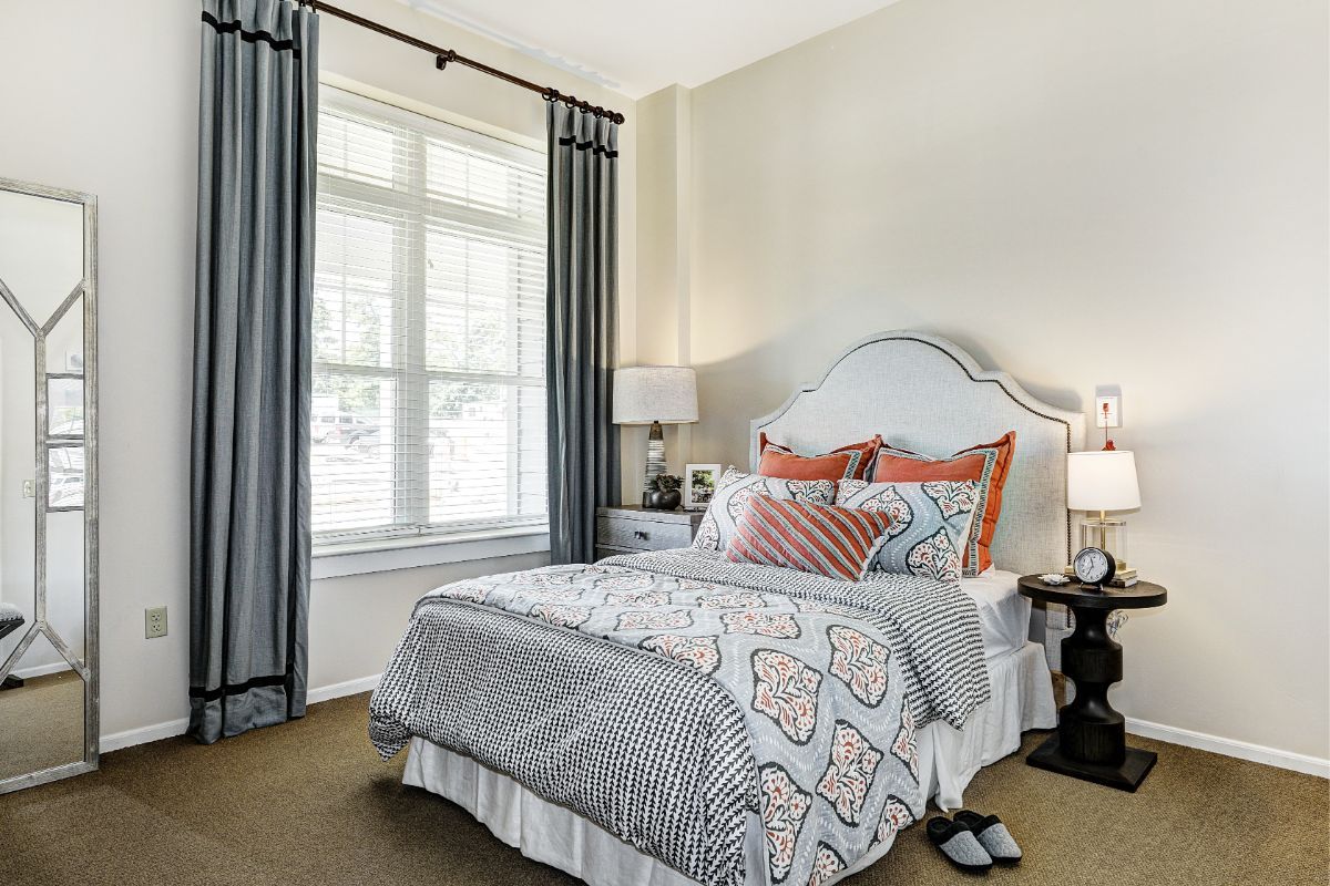 A cozy bedroom with a neatly made bed, decorative pillows, and natural light streaming through a large window.