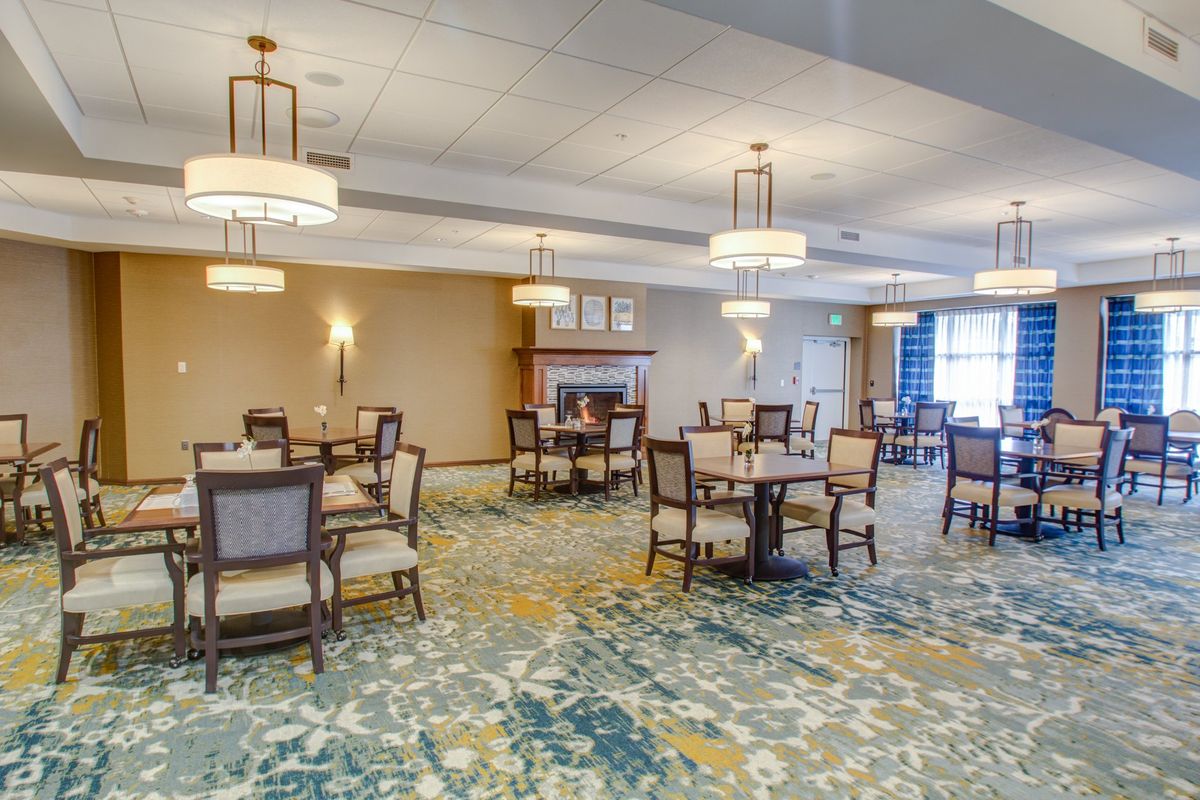 A well-lit dining area with elegantly arranged tables and chairs, featuring chandeliers and a cozy fireplace.