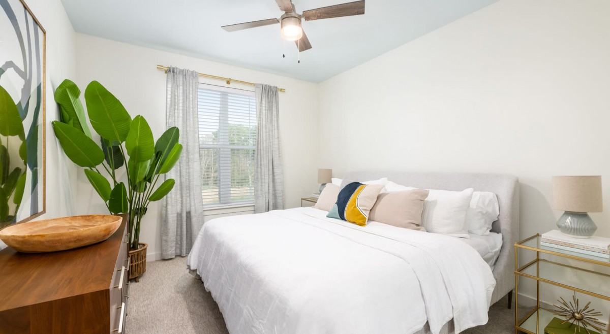 A bright and cozy bedroom featuring a large bed with decorative pillows, a ceiling fan, and a window with sheer curtains allowing natural light to enter.