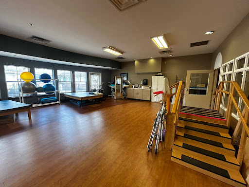 A spacious activity room equipped for rehabilitation and exercise, featuring wooden flooring and various equipment.