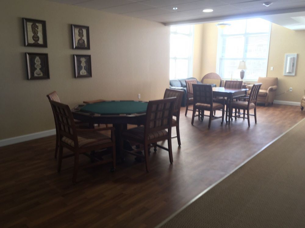A dining area with wooden floors, featuring several tables and chairs, illuminated by natural light from large windows.