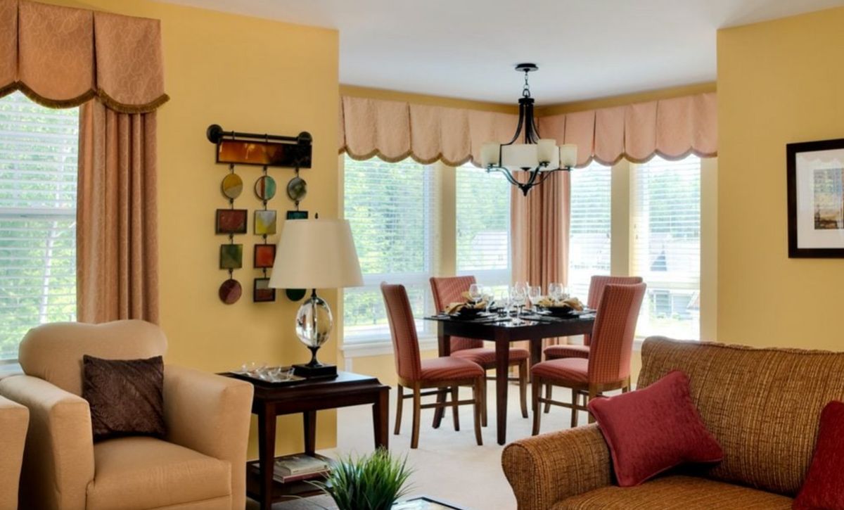 A cozy living room with an adjacent dining area, featuring a table set for four and decorated with warm tones and natural light from large windows.